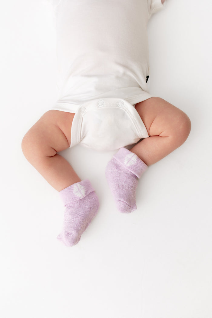 A baby lying on a white surface, wearing light purple knit socks with small white heart details and a white bodysuit.
