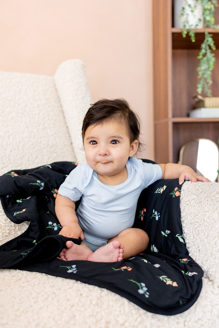 Baby sitting on a white sofa with Baby Blanket in Midnight Wildflower 1.0 print wearing a blue bodysuit