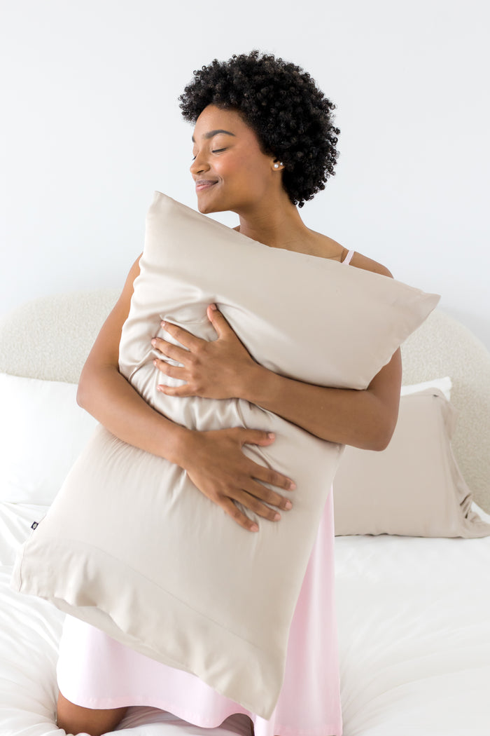 Female kneeling on a bed holding a  pillow with the Bamboo Sateen Pillowcase in Sand Stone on it