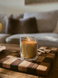 Busy Bees Classic Beeswax Jar Candle 12oz displayed on a wooden board with a lit flame and softly blurred living room background.