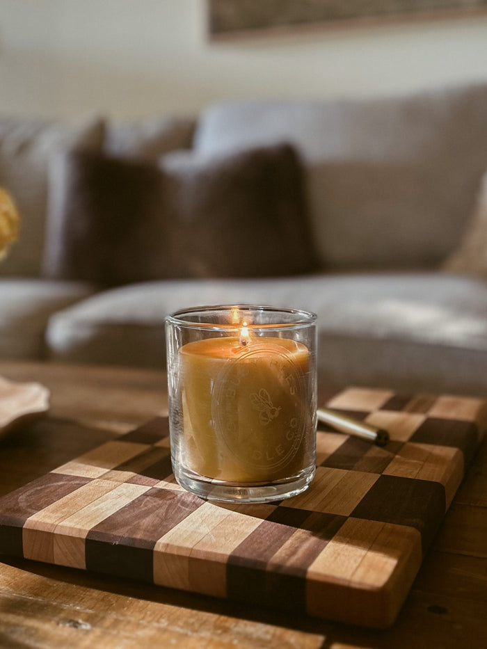 Busy Bees Classic Beeswax Jar Candle 12oz displayed on a wooden board with a lit flame and softly blurred living room background.