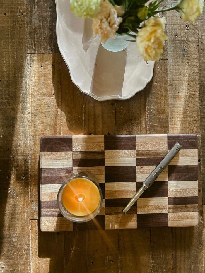 Lit Busy Bees Classic Beeswax Jar Candle 12oz displayed on a tiled surface beside a vase of flowers in warm ambient lighting.