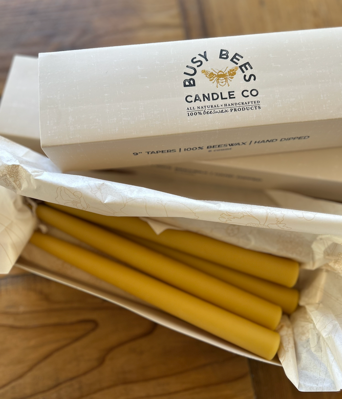 Busy Bees Hand Dipped 9 Inch Taper Candles 6 Pack partially wrapped in branded paper packaging on a wooden surface.