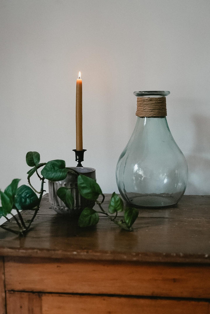 Single lit Busy Bees Hand Dipped 9 Inch Taper Candle displayed beside greenery and a glass bottle on a wooden cabinet.