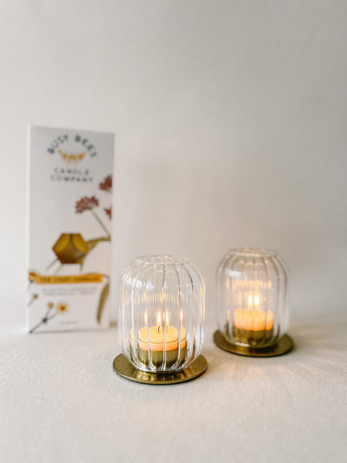 Busy Bees Tea Light Candles Box 10ct displayed beside two lit tea lights in ribbed glass holders against a minimal neutral background.