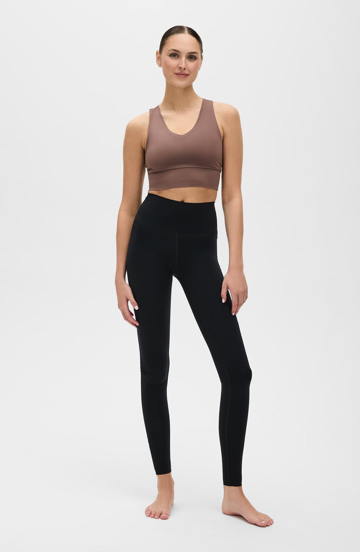 Woman wearing an Elastique Athletics Adorn brown sports bra and black leggings on a white background