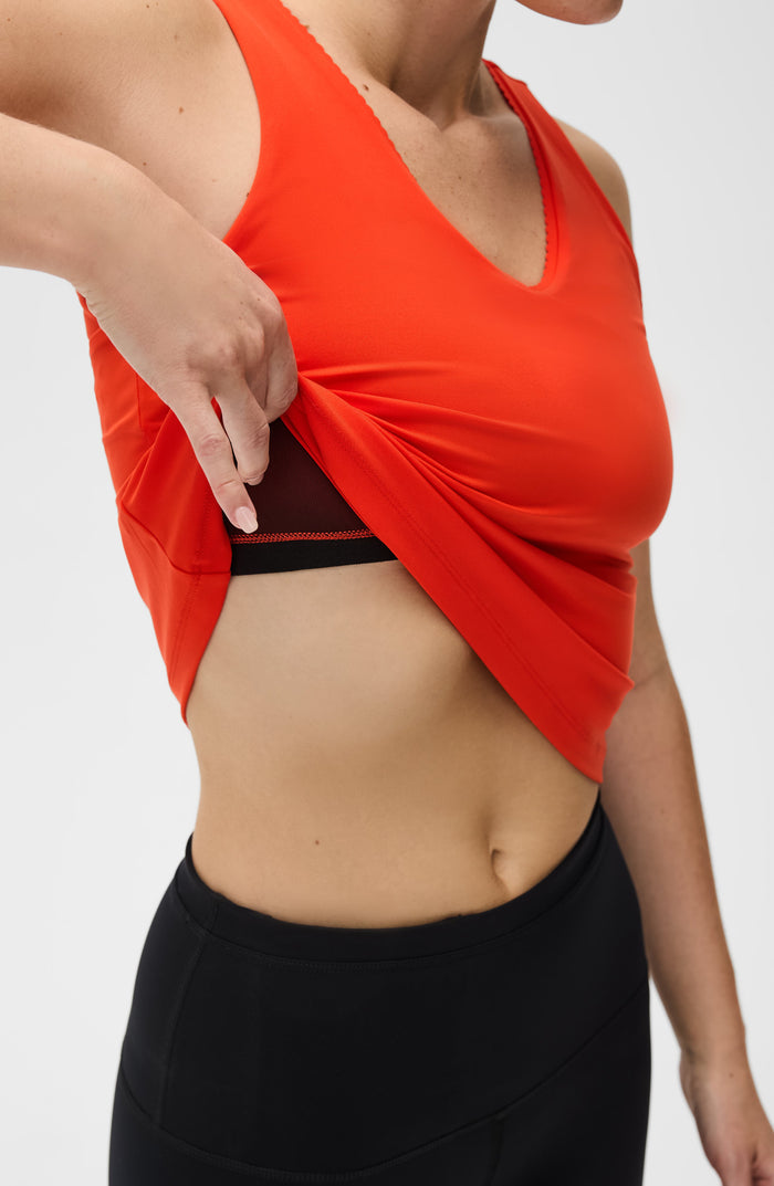 Close up detail of a woman lifting the hem of the Elastique Athletics Adorn Tank in bright orange paired with black leggings.