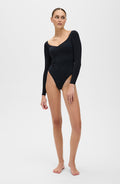 Woman standing barefoot in a studio while wearing the Elastique Athletics Iconic Compression Long Sleeve Bodysuit Thong in black with a scoop neckline and fitted silhouette.