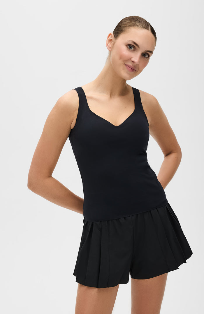 Woman smiling while wearing the Elastique Athletics Iconic MicroPerle Tank in black paired with a pleated athletic skirt.