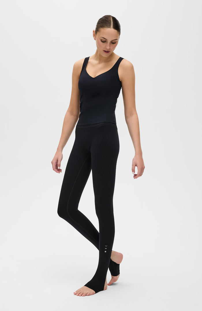 Woman walking barefoot in a studio while wearing the Elastique Athletics Iconic MicroPerle Tank in black with fitted leggings.