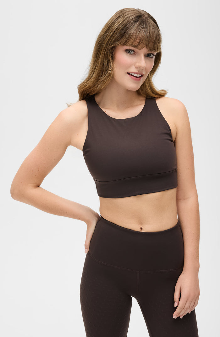 Woman posing in the Elastique Athletics L'Original dark brown sports bra and black leggings in a minimal studio setting.