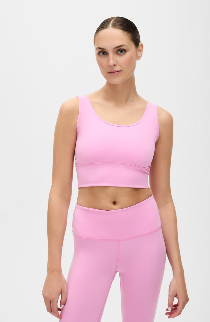 Woman standing barefoot while wearing the Elastique Athletics Rise Bra Vie en Rose in soft pink with matching high waisted leggings against a neutral studio background.