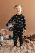 Toddler wearing Black Long Sleeve Jogger Set in Monster Truck print holding a truck standing on sand and rock setup