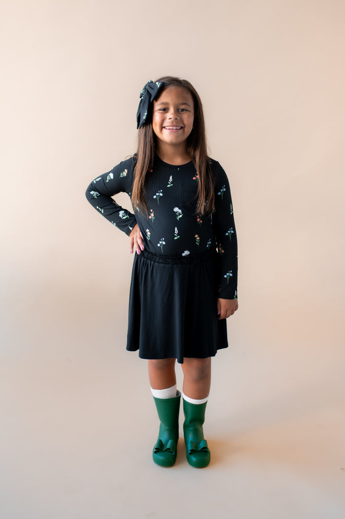 Girl wearing Long Sleeve Toddler Crew Neck Tee in Midnight Wildflower print over black skort and green shoes along with a similar print large alligator clip bow on a beige background