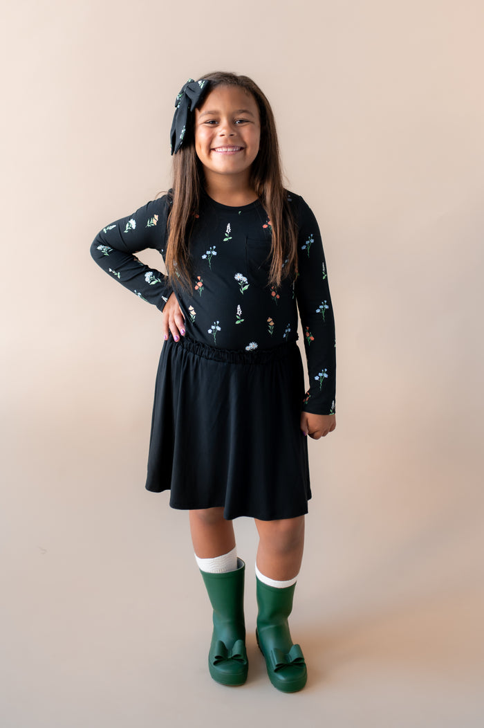 Girl posing in Long Sleeve Toddler Crew Neck Tee in Midnight Wildflower print over black skort and green shoes along with a similar print large alligator clip bow on a beige background