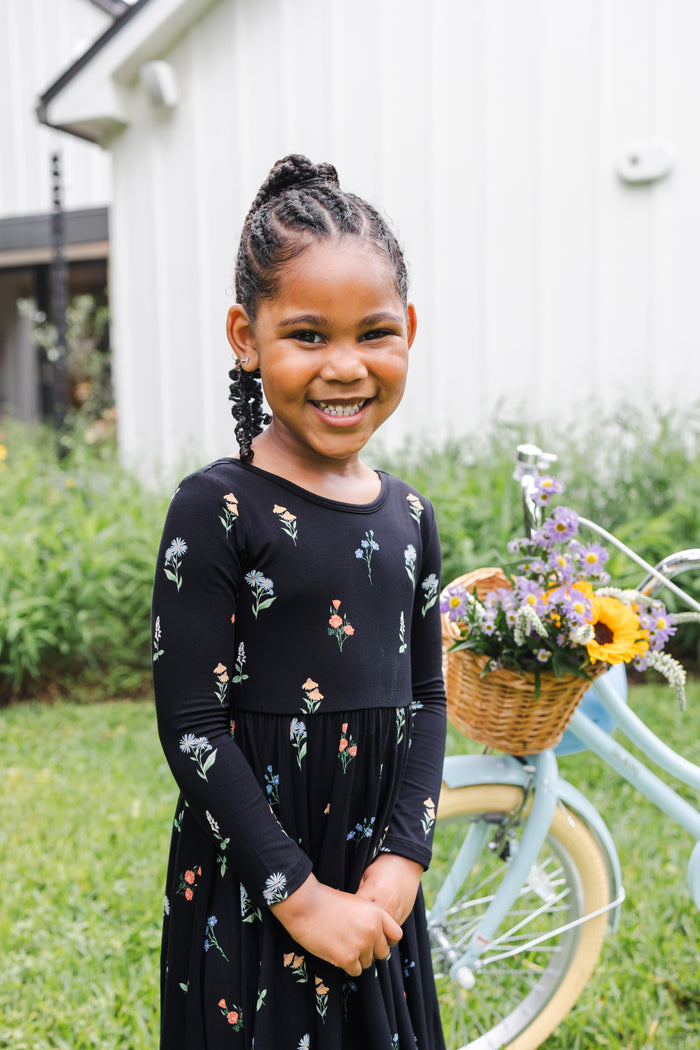 Girl smiling outdoors with a bicycle in grass wearing Long Sleeve Twirl Dress in Midnight Wildflower print
