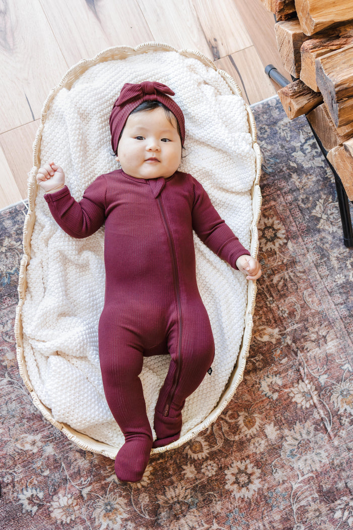 Baby wearing Ribbed Zipper Footie and Ribbed Headwrap in Burgundy lying in a basket