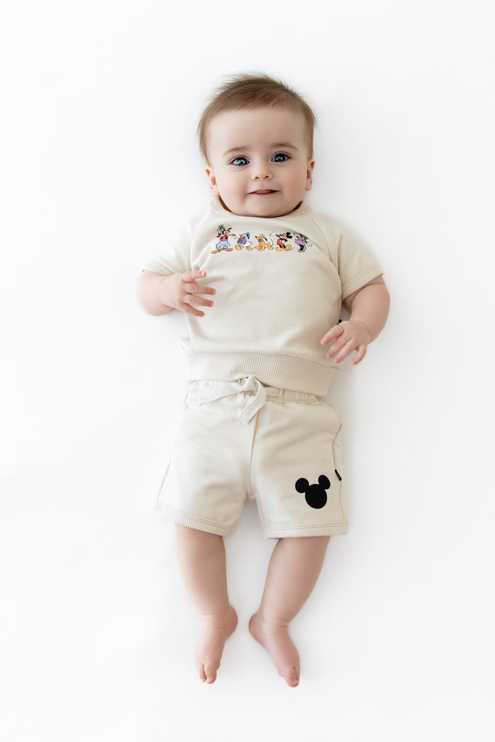 Baby wearing a cream colored matching set with mickey friends on the shirt and black mickey head on the short