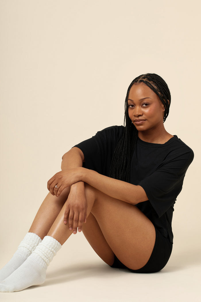 Woman sitting on a beige background wearing a black outfit and white socks.