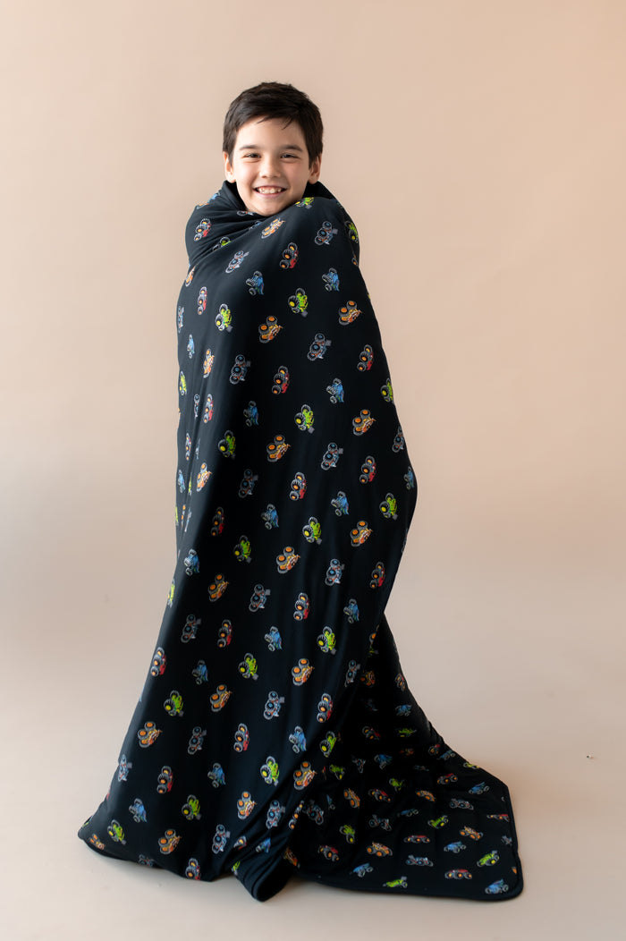 Boy wrapped in black Youth Blanket in Monster Truck print on beige background