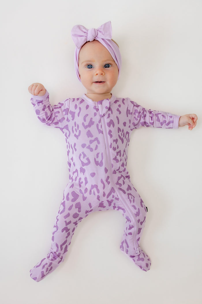 Baby wearing zippered footie in Thistle Leopard print with a thistle bow headband on a beige background